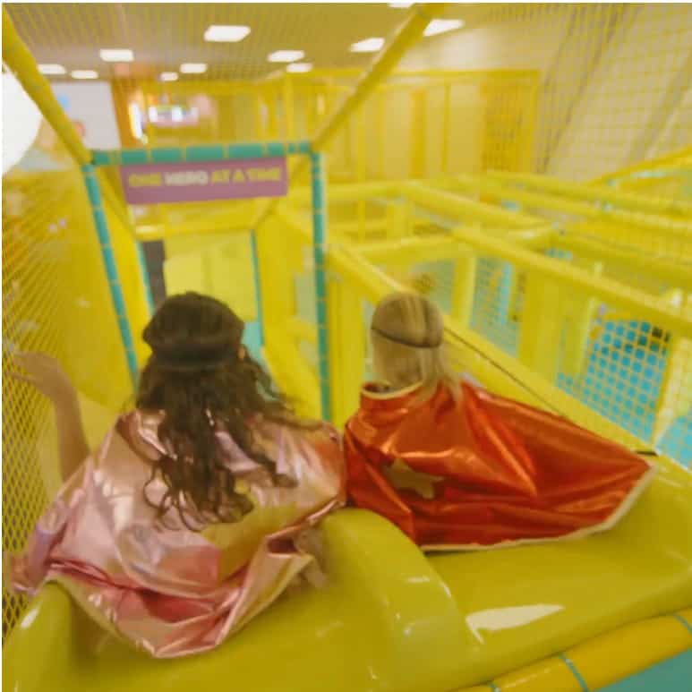 Dos niños con capas y cintas para la cabeza están sentados en lo alto de un tobogán amarillo dentro del área de juegos de superhéroes de Chuck E. Cheese.