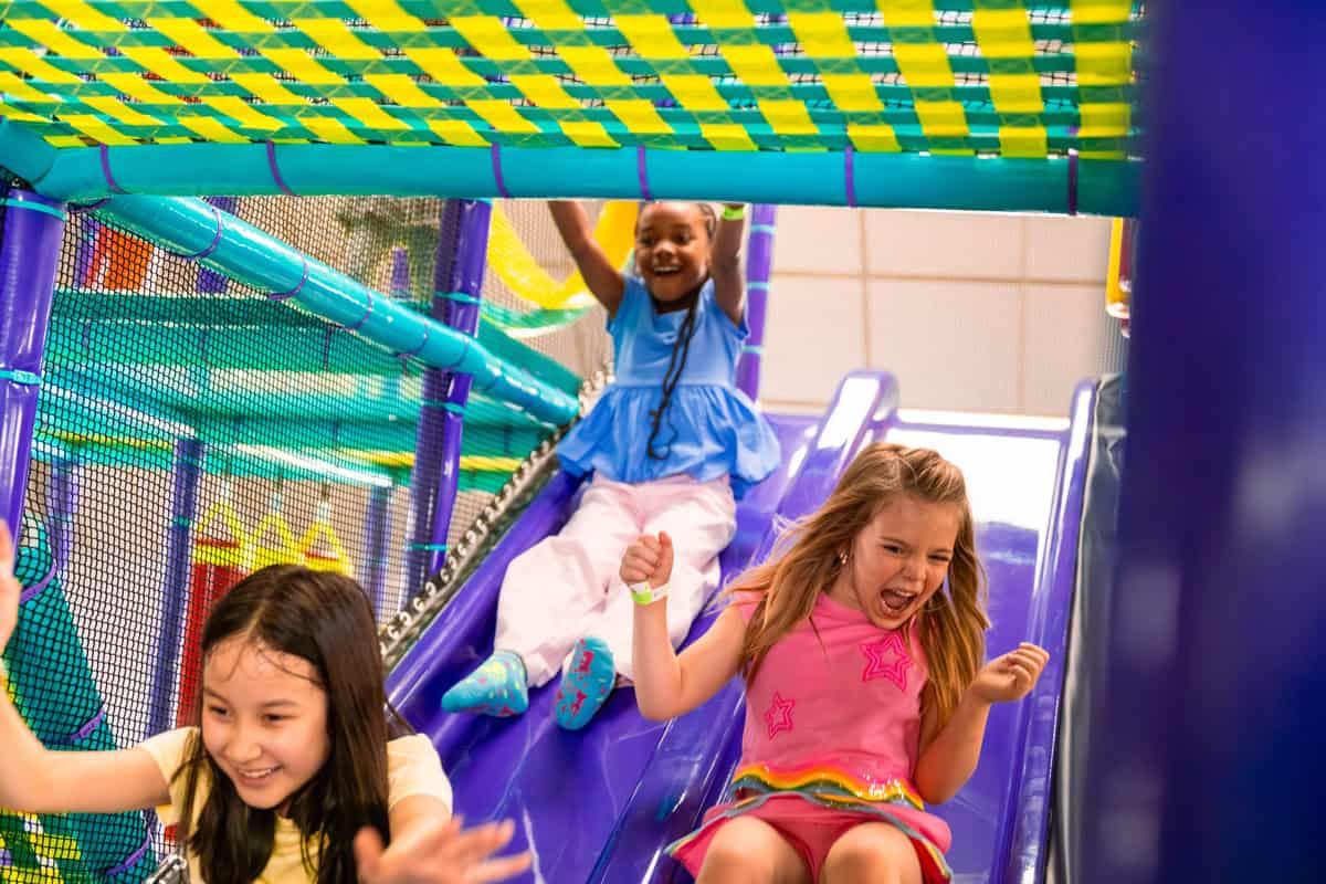 Tres chicas riendo y deslizándose por un tobogán morado en la Zona de Aventuras de Chuck E. Cheese.